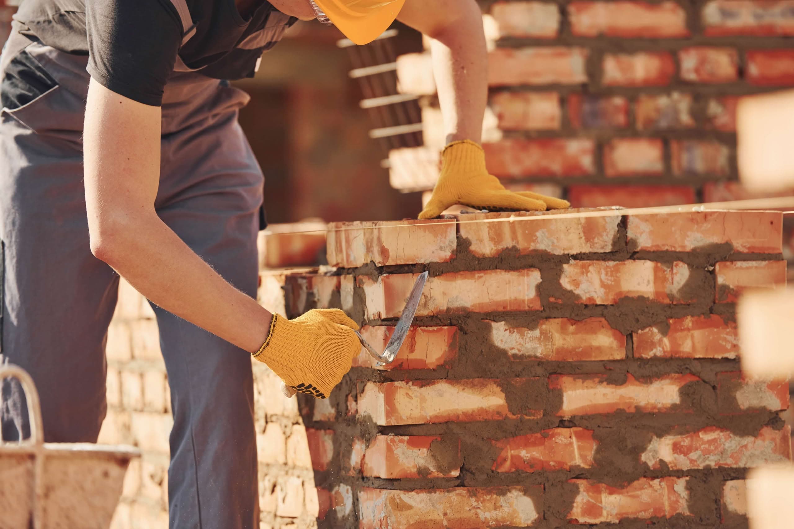 Construction worker in a yellow hard hat and safety gloves using a trowel to apply mortar while laying a red brick wall on a sunny building site.