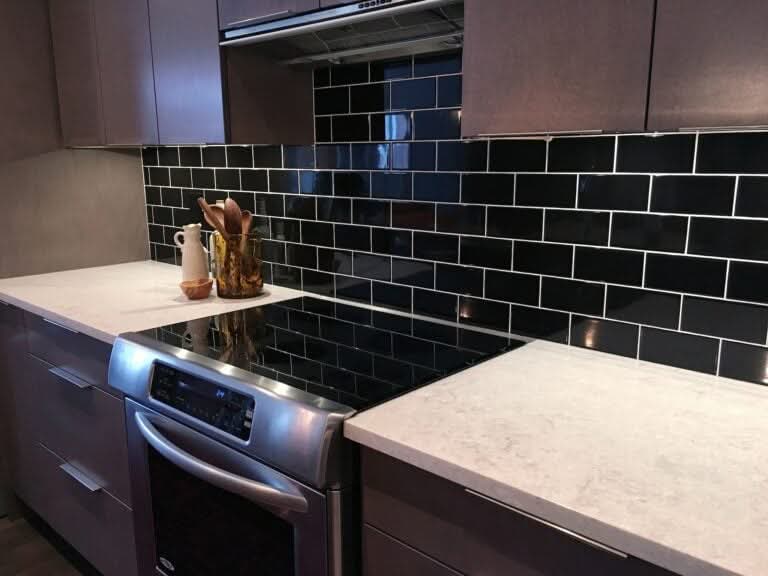 A sleek kitchen featuring a glossy black subway tile backsplash with white grout, paired with white quartz countertops and a stainless steel electric range.
