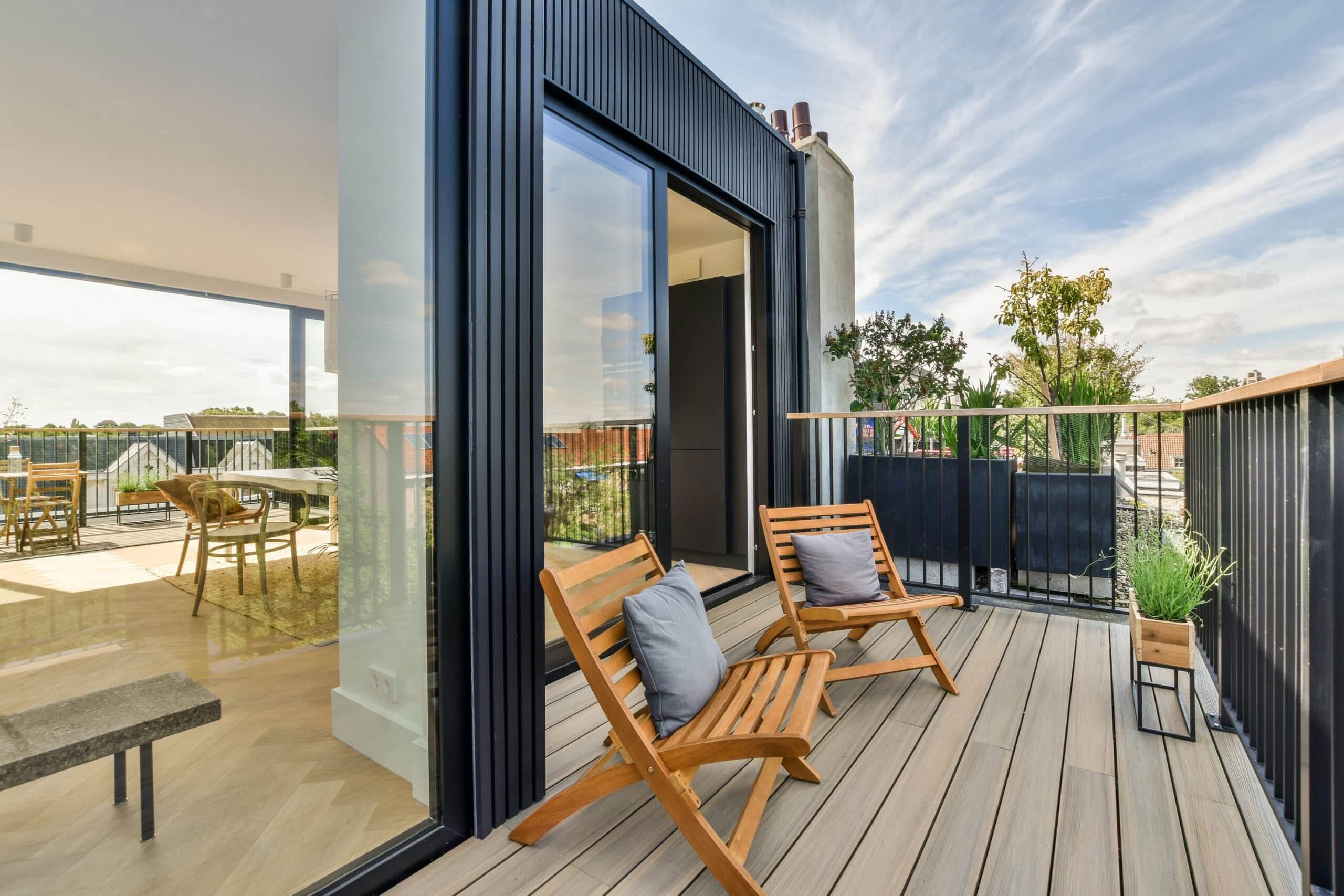 A modern rooftop balcony featuring light gray wooden decking, two wooden folding chairs with gray pillows, and black metal railings with a view into a sunlit apartment through a sliding glass door.