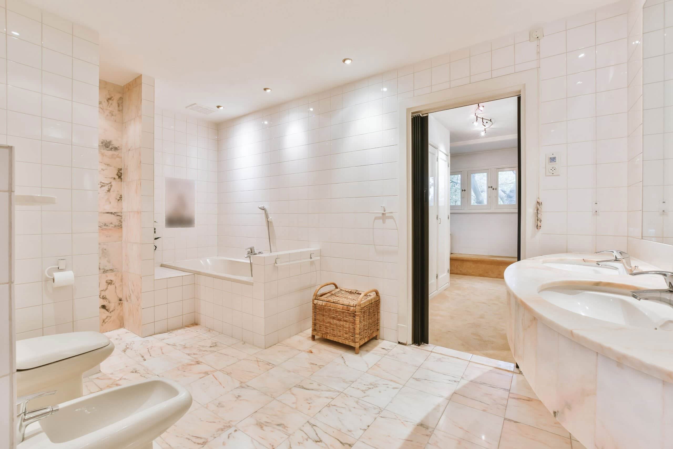 Spacious modern bathroom featuring white tile walls, pink-veined marble floors, a built-in bathtub, and a curved double marble vanity.