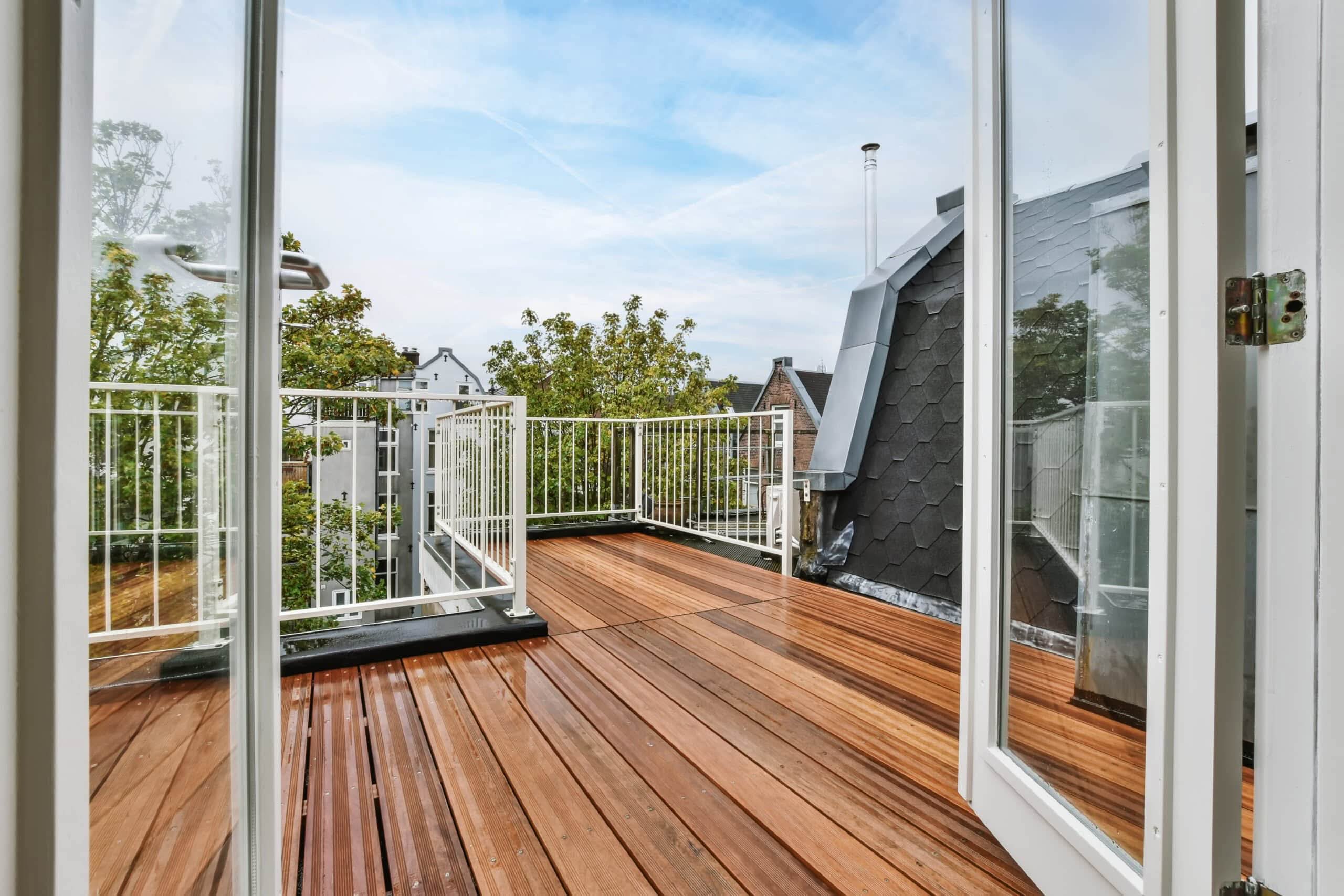 A view through open white French doors onto a modern rooftop deck with wet reddish-brown wood planks, white metal railings, and a view of surrounding city buildings and trees.