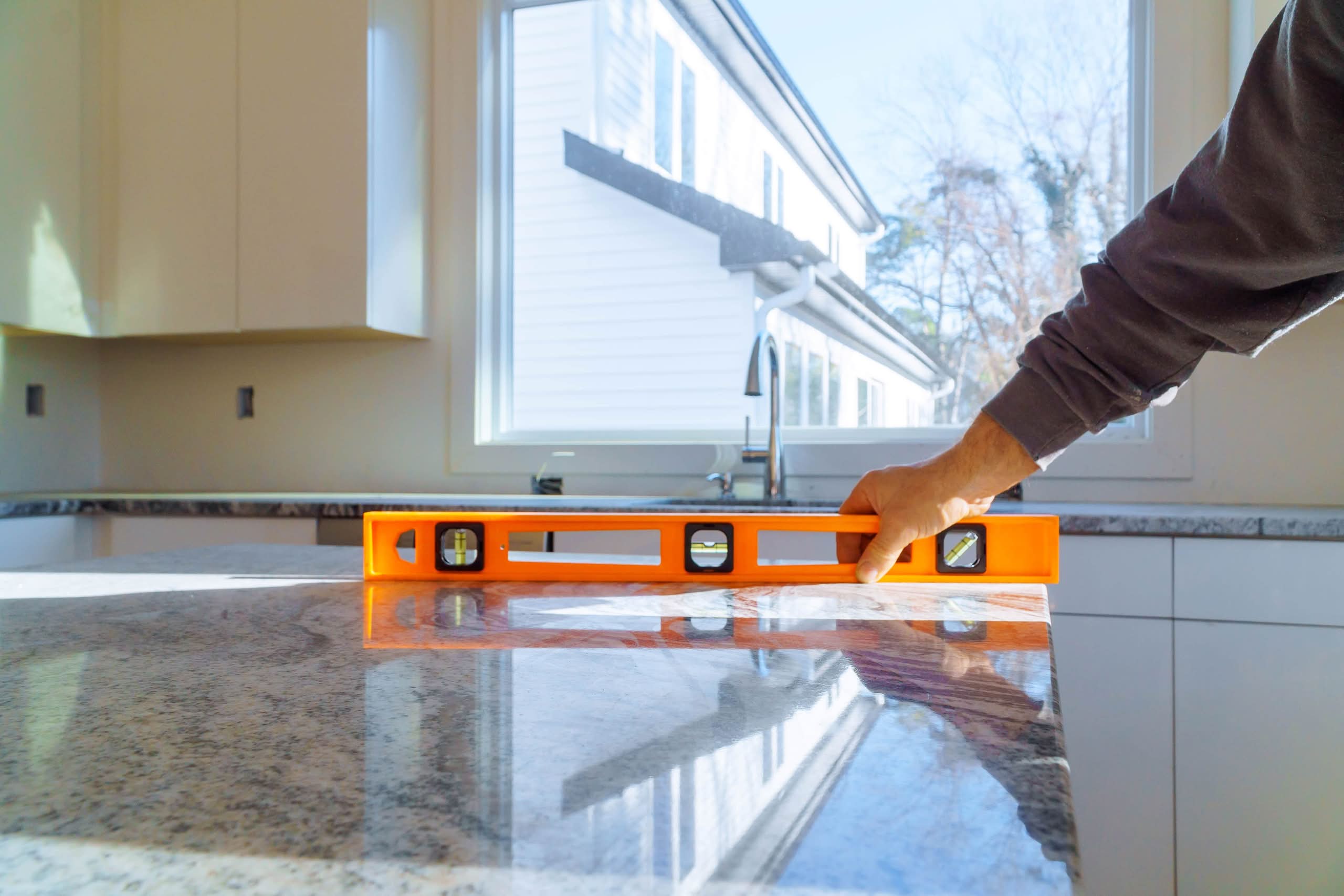 Close-up of a contractor's hand using an orange spirit level on a polished granite kitchen island countertop during installation, with a bright window and white kitchen cabinetry in the background.