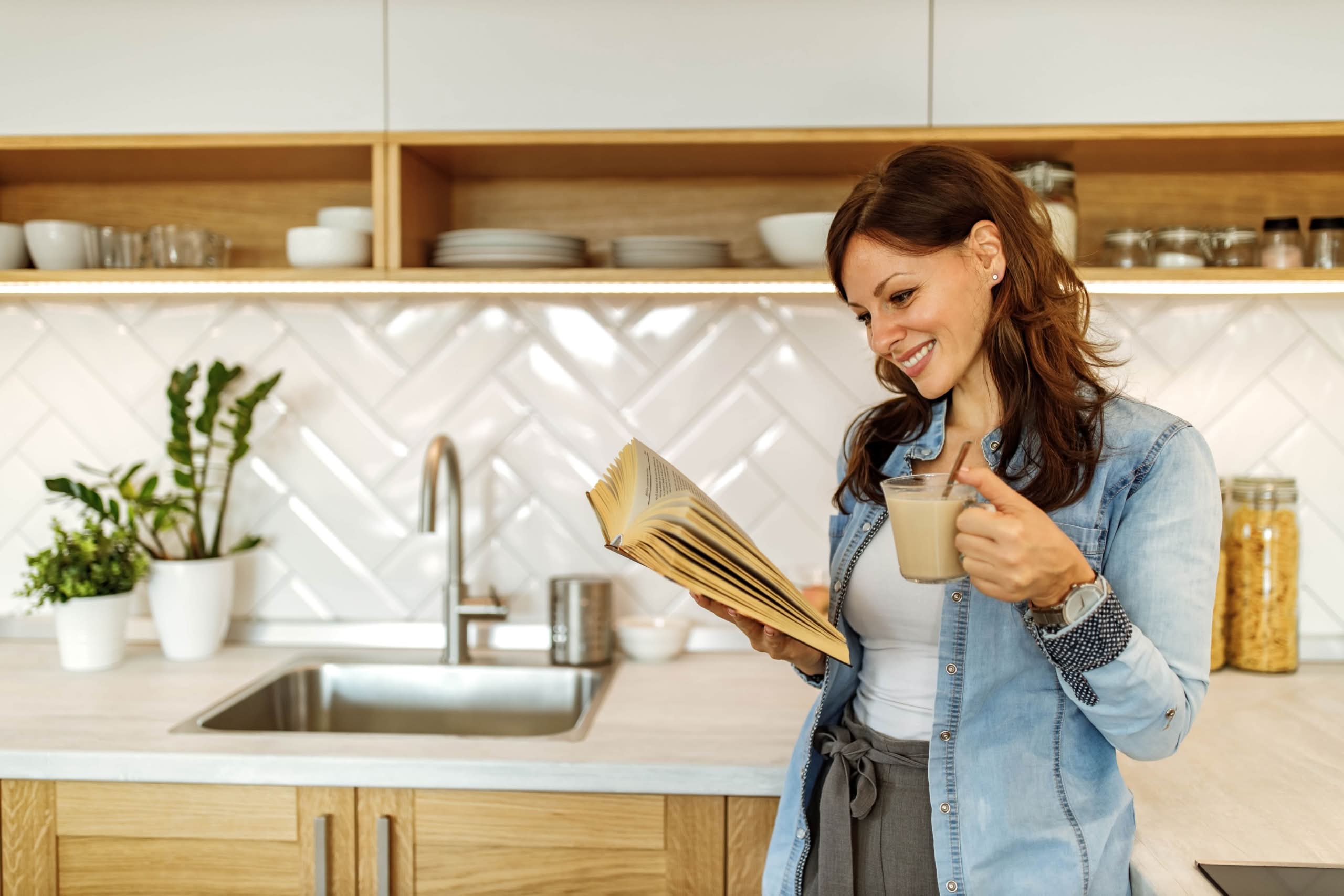 A smiling woman in a denim shirt holding a cup of coffee and reading a cookbook in a bright, modern kitchen with white herringbone tile backsplash and wooden cabinetry.