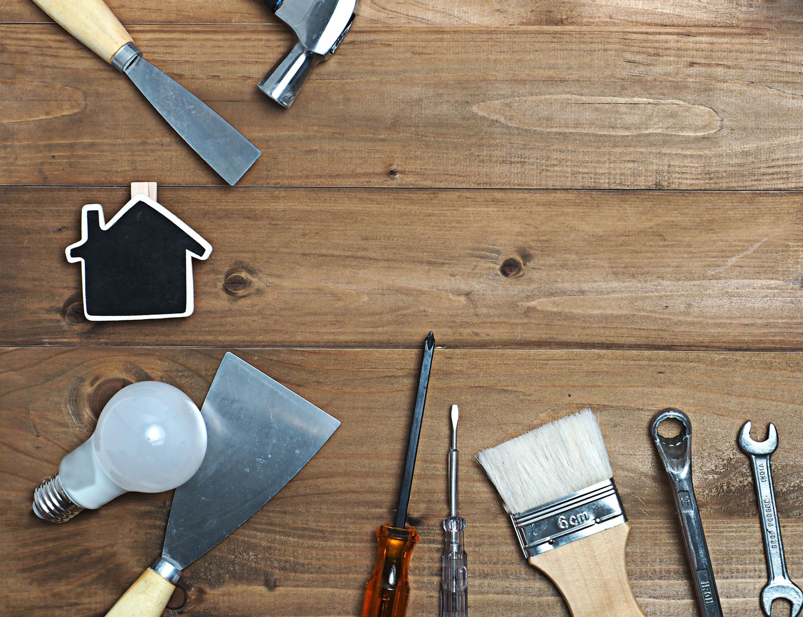 Handyman for small home repairs and honey-do lists 1 A top-down view of various hand tools including a paintbrush, screwdrivers, wrenches, a putty knife, and a lightbulb arranged on a wooden surface around a small house-shaped chalkboard.