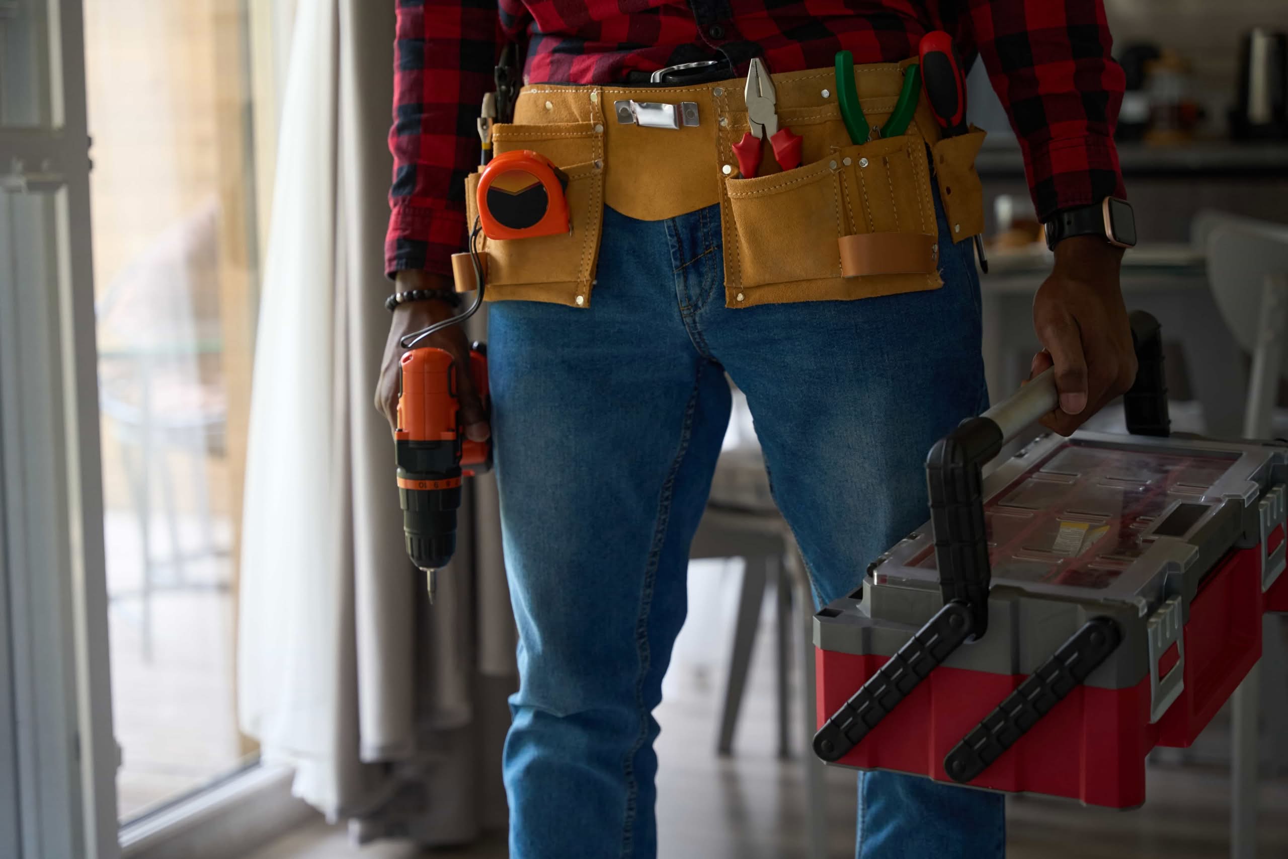 A professional handyman repair services in Atlanta, GA wearing a tool belt and holding a power drill and toolbox indoors.