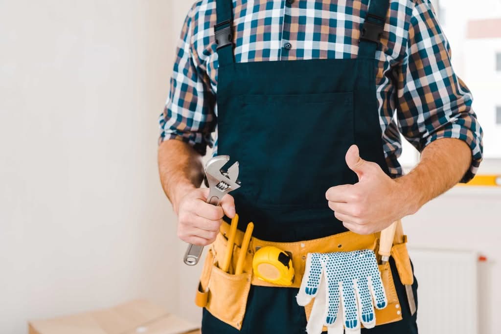 A professional handyman in Charlotte, NC wearing overalls and a tool belt, holding a wrench and giving a thumbs up.