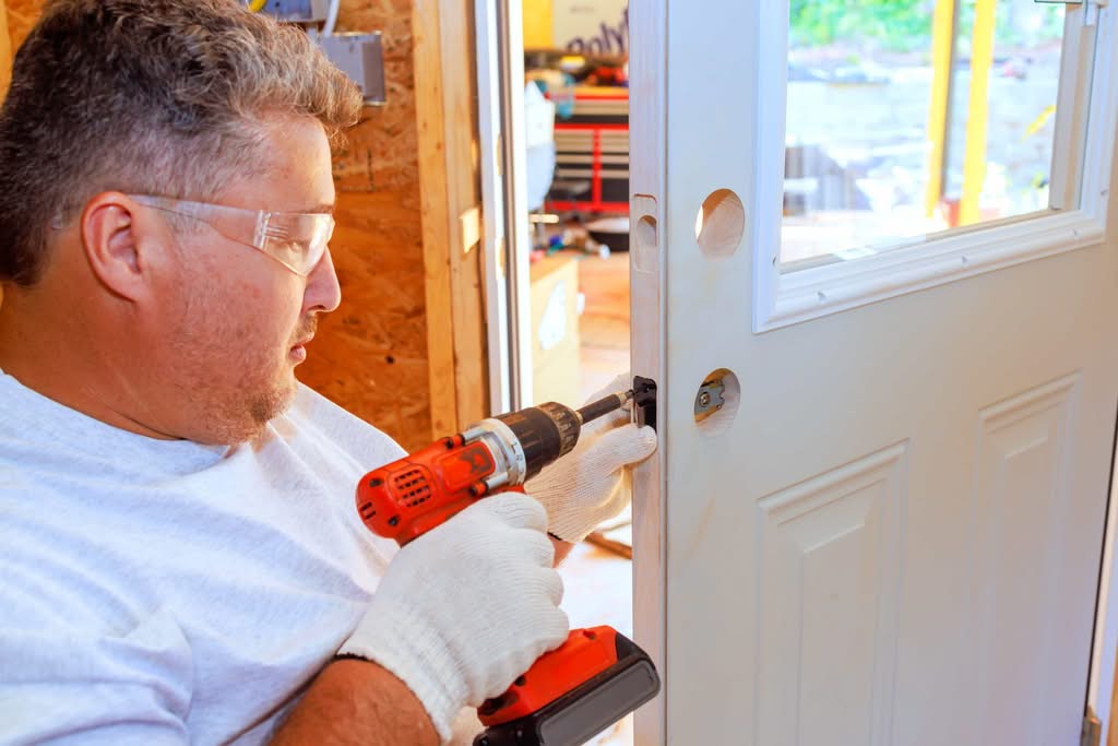 A professional locksmith wearing safety glasses and gloves uses a power drill to install a door lock in Las Vegas, NV.